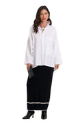 A woman modeling a 100% cotton white long-sleeve button-up shirt with patch pockets, paired with a black maxi skirt featuring a white stripe near the hem and black ankle boots, against a plain white background.