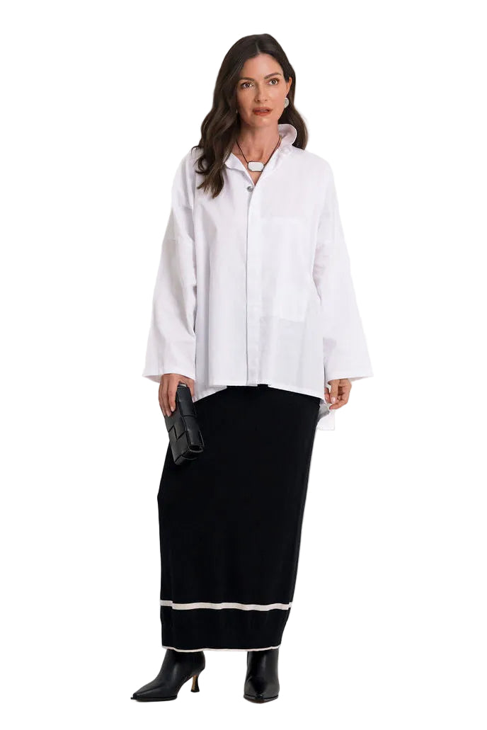 A woman modeling a 100% cotton white long-sleeve button-up shirt with patch pockets, paired with a black maxi skirt featuring a white stripe near the hem and black ankle boots, against a plain white background.