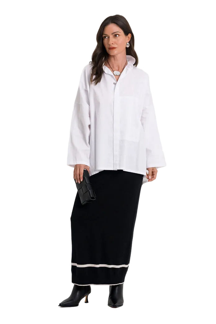 A woman stands against a plain light gray studio background wearing a 100% cotton loose white long-sleeve button-up shirt and a long black skirt with a white stripe near the hem, paired with black ankle boots.