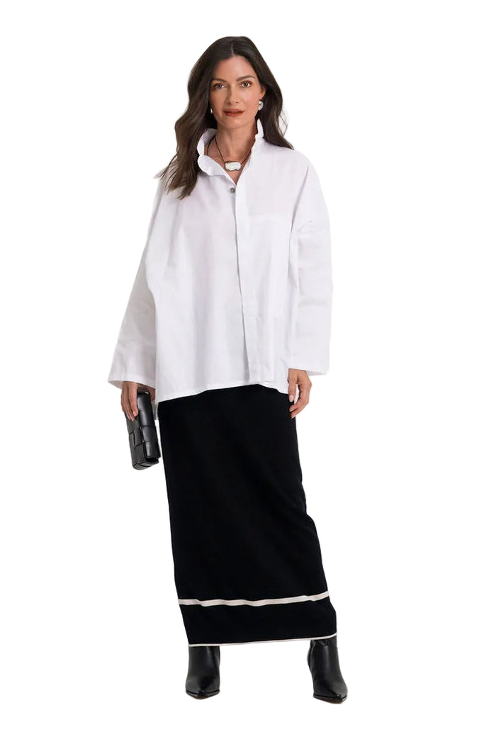 A woman modeling a 100% cotton white long-sleeve button-up shirt paired with a black maxi skirt featuring a white stripe near the hem, and black ankle boots, against a plain white studio background.