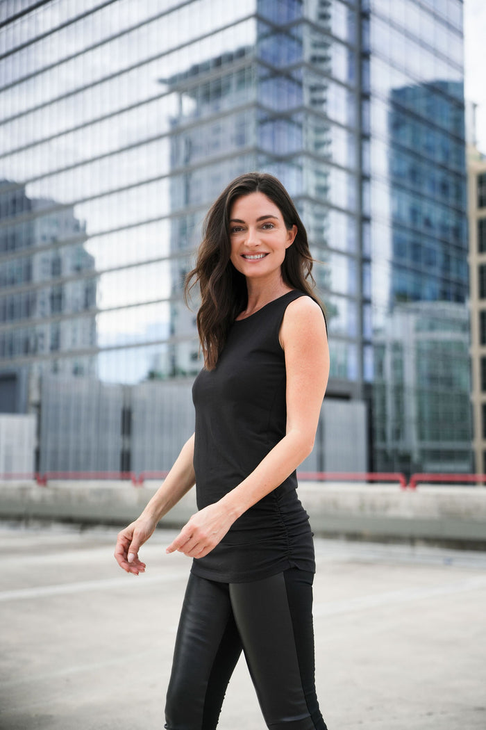 Woman in black sleeveless ruche top and vegan leather leggings standing in front of modern glass buildings.