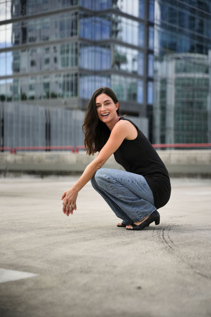 Woman squatting on a street wearing black lycra cotton ruche shirt and jeans with modern buildings in the background