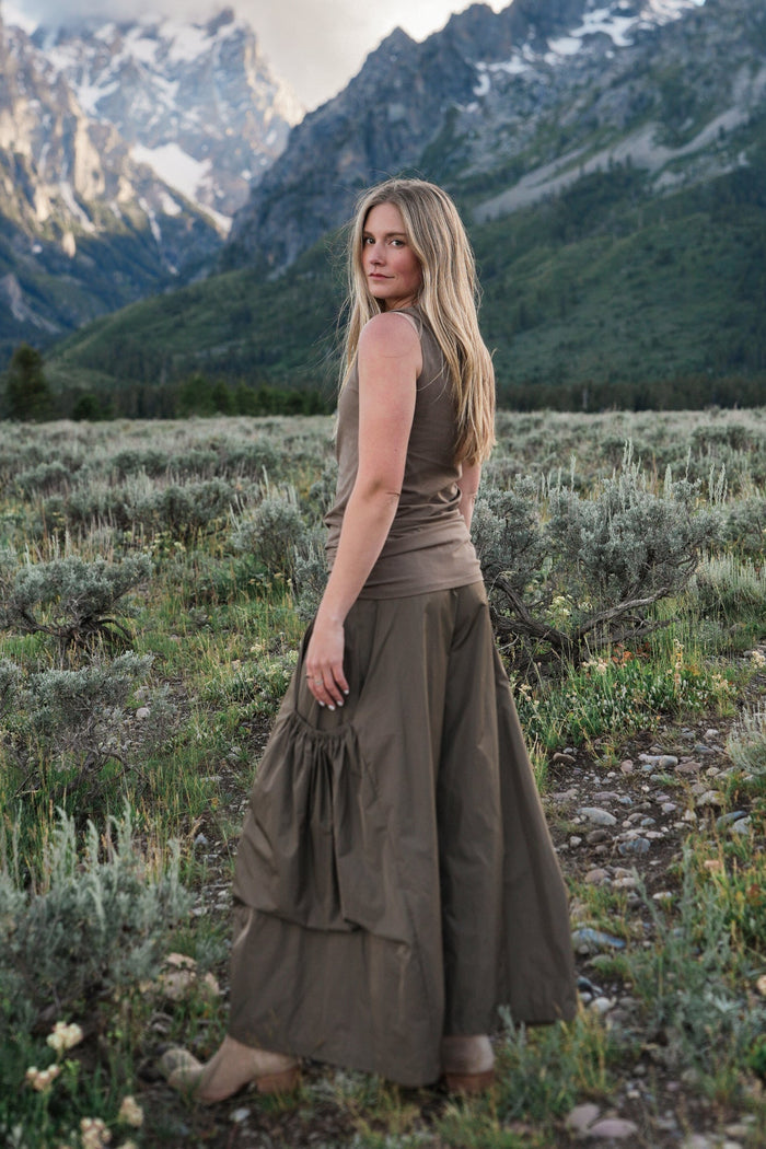 A woman stands in a grassy mountain meadow with snow-capped peaks and a blue sky behind her, wearing a sleeveless taupe-brown cotton lycra tank top and tan boots.