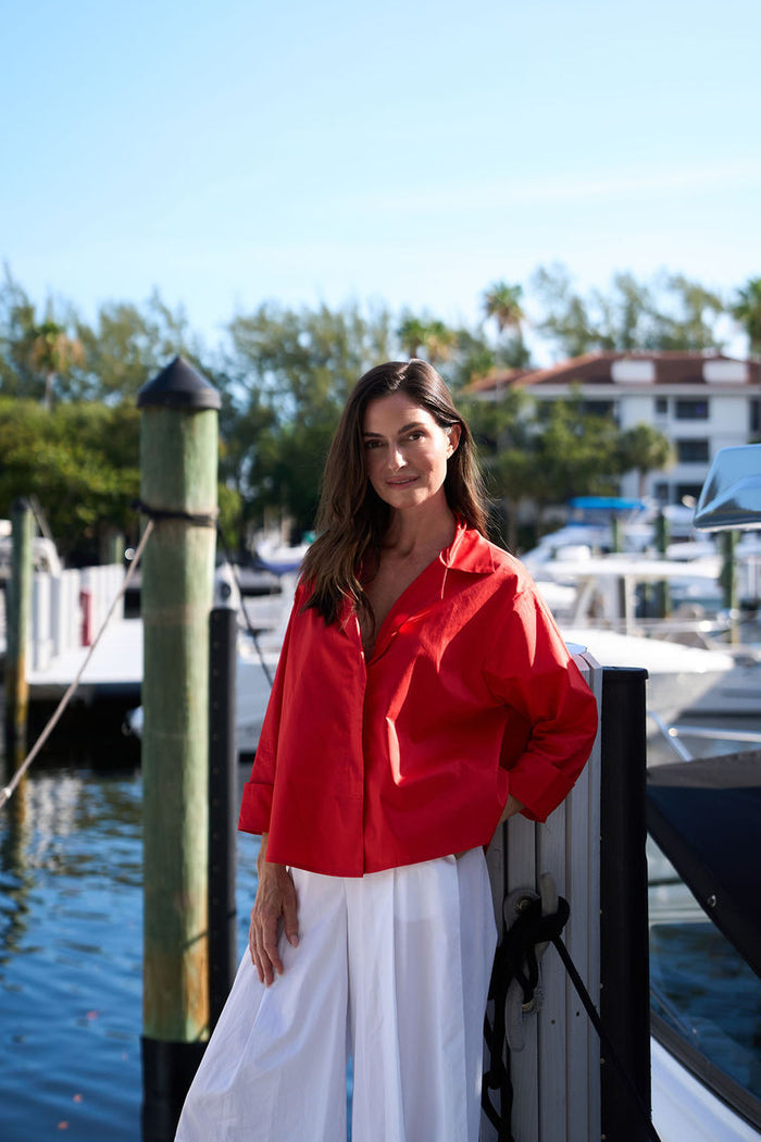 Woman in a red cotton shirt and white cotton pants standing by a dock with boats in the background