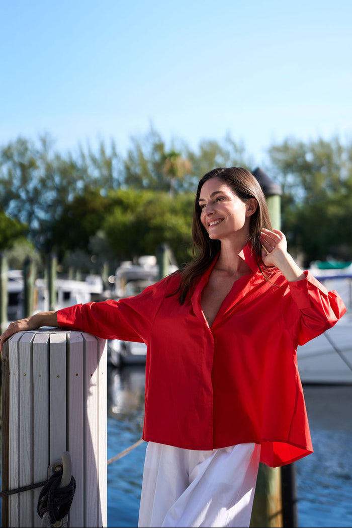 Woman in a red cotton blouse standing by a dock with boats and greenery in the background
