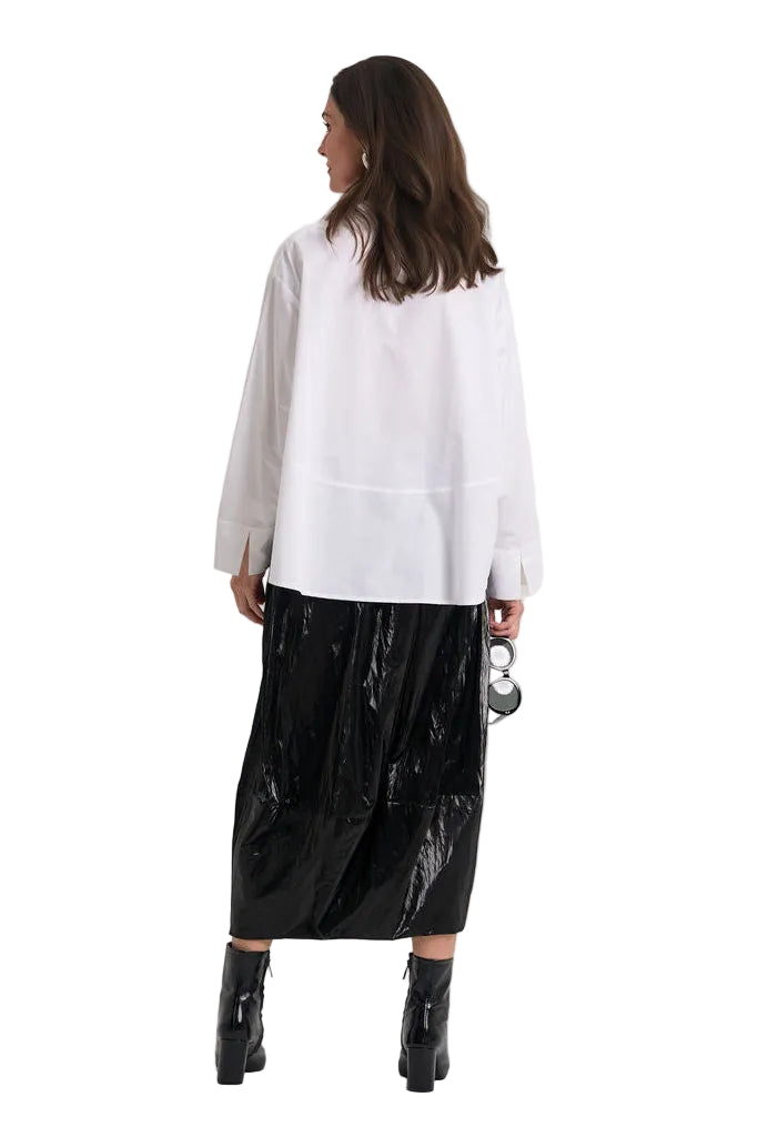 A woman shown from the back wearing a loose white 100% cotton long-sleeve top and a shiny black midi skirt, standing against a plain light background.