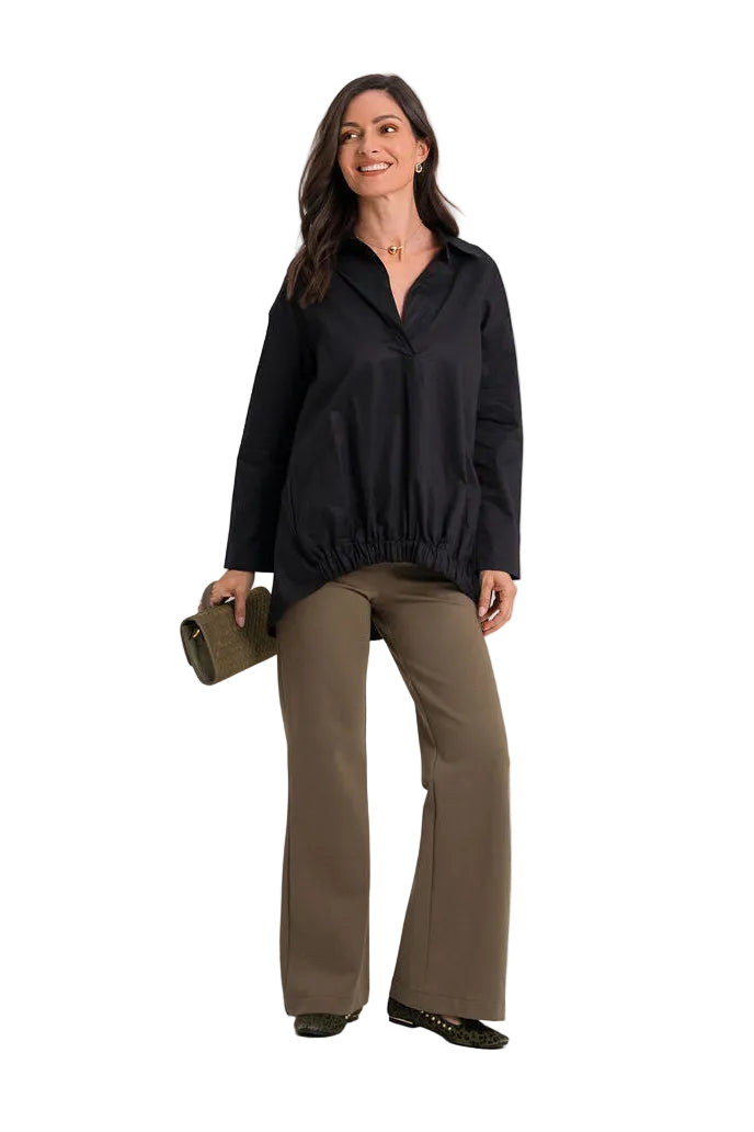 A woman modeling a 100% cotton black long-sleeve collared jacket with a gathered elastic hem, paired with olive green wide-leg pants and holding a small dark clutch, against a plain white studio background.