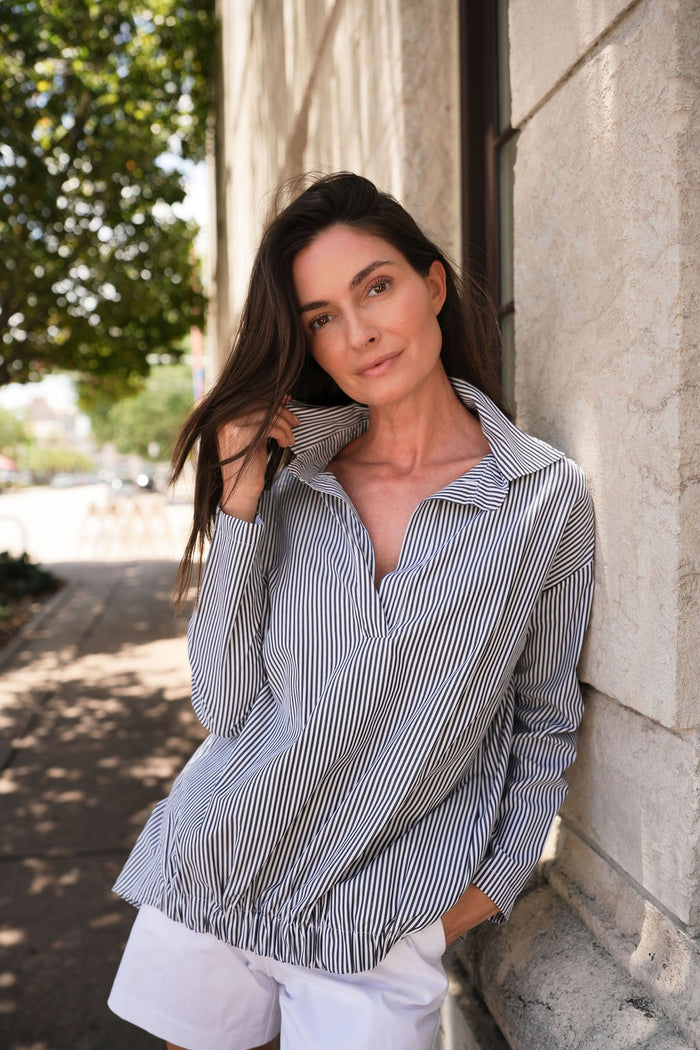 Woman leaning against a stone wall wearing a PLANET striped 100% cotton shirt and white shorts.