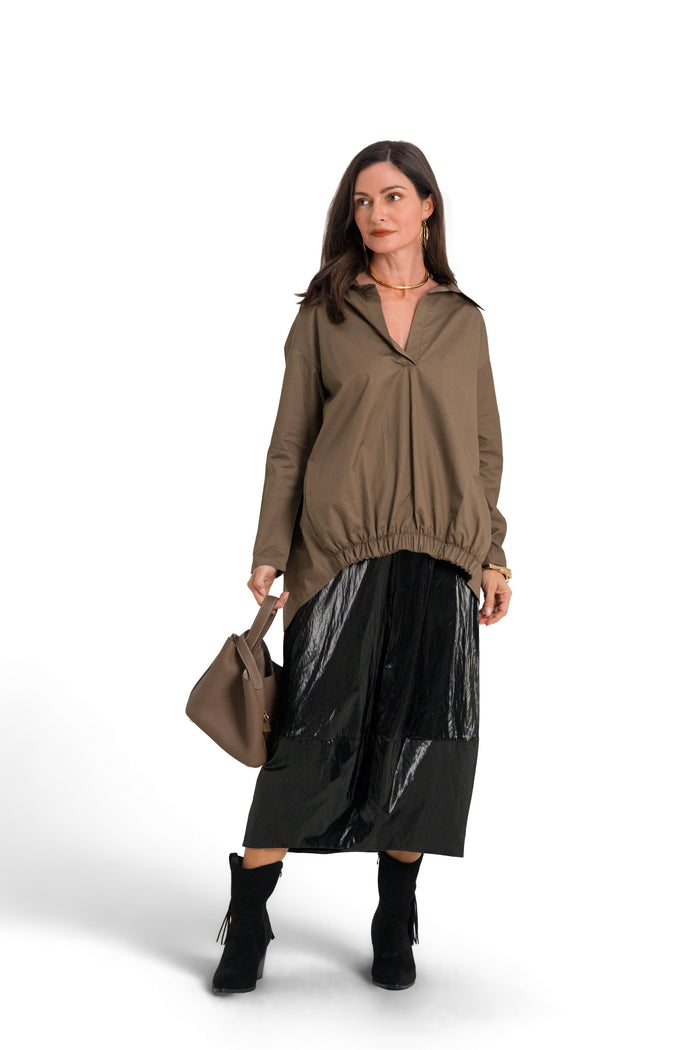 A woman stands against a white studio background wearing a 100% cotton brownish olive long-sleeve top, a shiny black skirt, black ankle boots, and holding a brown handbag.