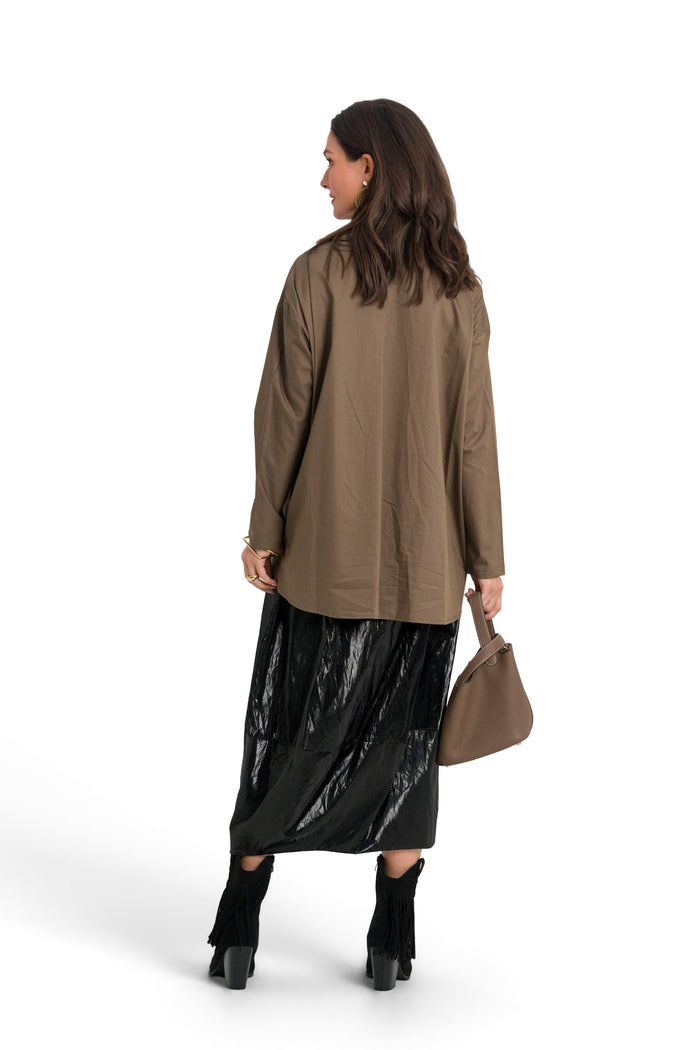 A woman is shown from the back wearing an 100% cotton oversized taupe-brown long-sleeve top, a shiny black midi skirt, black ankle boots, and holding a brown handbag against a solid white background.