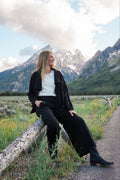 A woman wearing a black jacket, white top, black pants, and black boots sits on a log in a grassy field with mountains and a cloudy sky in the background.