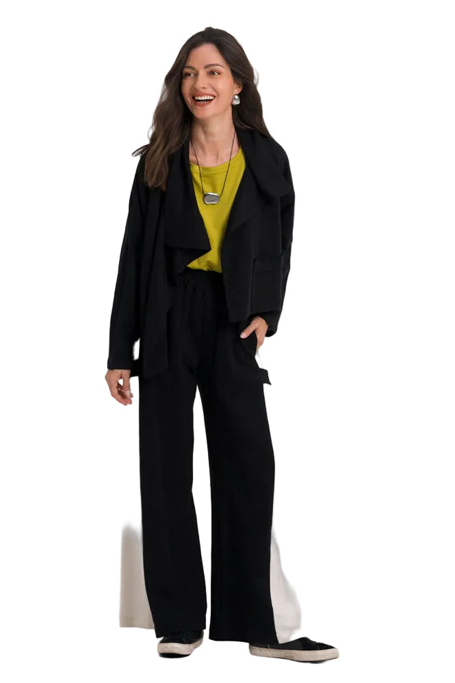 A woman modeling a black wide-leg pantsuit with a 100% cotton black blazer, worn over a bright yellow-green top, standing against a solid white studio background.