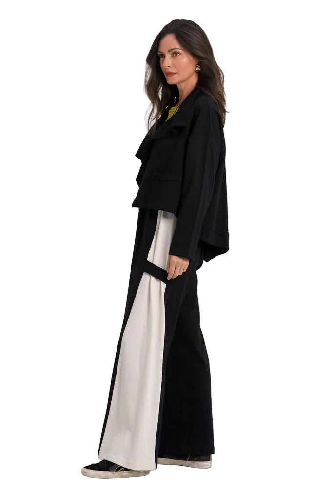 A woman stands in profile wearing a long 100% cotton black draped jacket or coat over wide-leg black pants with bold white side panels, photographed against a clean white studio background.