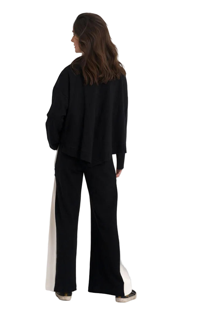 A woman is shown from behind wearing a 100% cotton black long-sleeve jacket and black wide-leg pants with white side panels, standing against a plain white studio background.