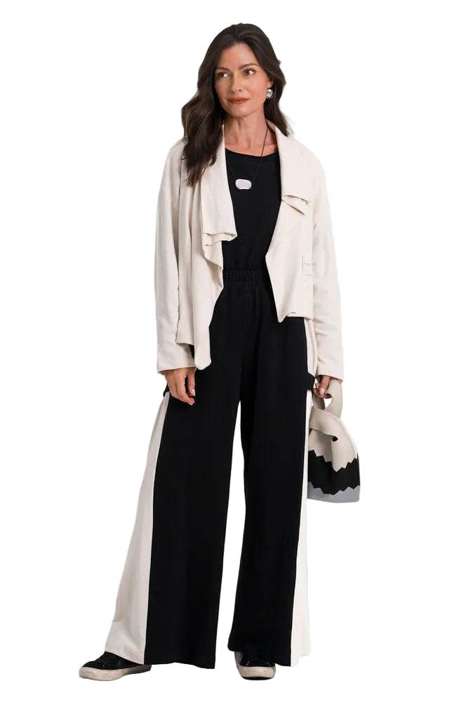 A woman stands against a plain white studio background wearing a black wide-leg jumpsuit with a 100% cotton white side panel, a cream open-front jacket, black shoes, and holding a small black-and-white handbag.