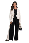 A woman stands against a plain white studio background wearing a black wide-leg jumpsuit with a 100% cotton white side panel, a cream open-front jacket, black shoes, and holding a small black-and-white handbag.