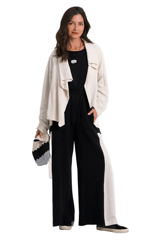 A woman stands against a plain white studio background wearing a black wide-leg jumpsuit with side pockets, a light 100% cotton beige open-front cardigan, and holding a small black and white patterned handbag.