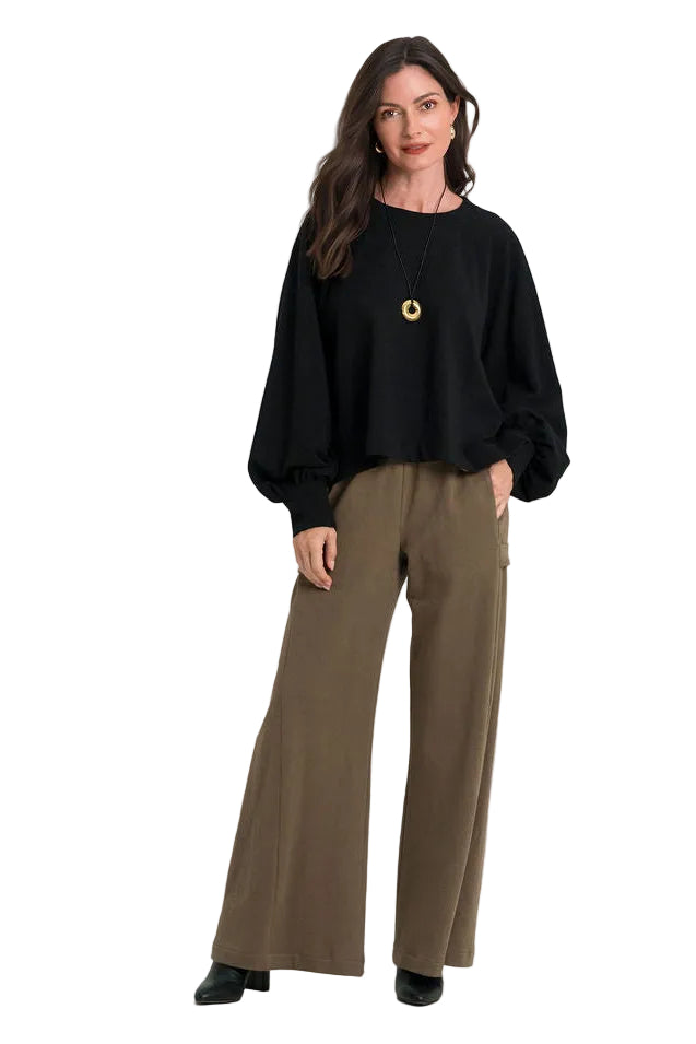 A woman modeling a 100% cotton black long-sleeve top with dramatic balloon sleeves and wide-leg brown trousers, standing against a plain white studio background.