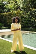 A woman with curly black hair stands outdoors beside a swimming pool on green grass, wearing a matching pale yellow collared top and 100% linen wide-leg pants set.