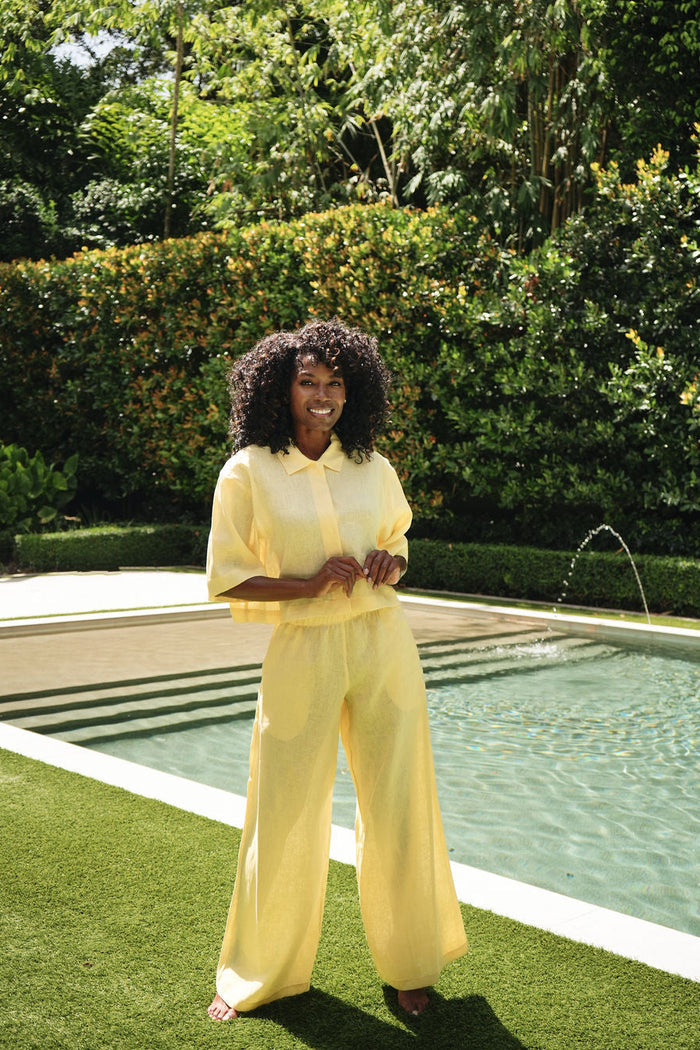A woman with curly black hair stands outdoors beside a swimming pool on green grass, wearing a matching pale yellow collared top and 100% linen wide-leg pants set.