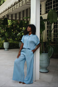 A woman wearing a light blue short-sleeve collared top and matching 100% linen wide-leg pants stands on a covered patio beside a white column, with large potted plants and a house exterior in the background.