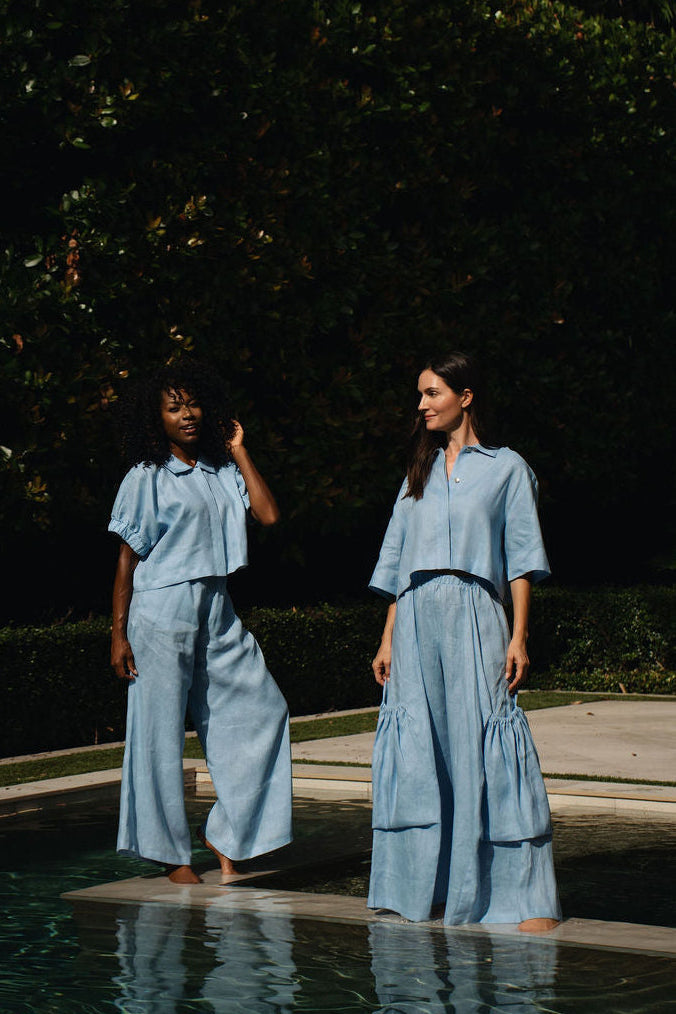 Two women wearing matching light blue 100% linen two-piece sets stand barefoot in a swimming pool at night, with dark green hedges in the background.