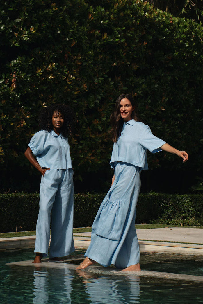 Two women modeling matching 100% linen light blue two-piece outfits by a swimming pool, with a lush green garden background.