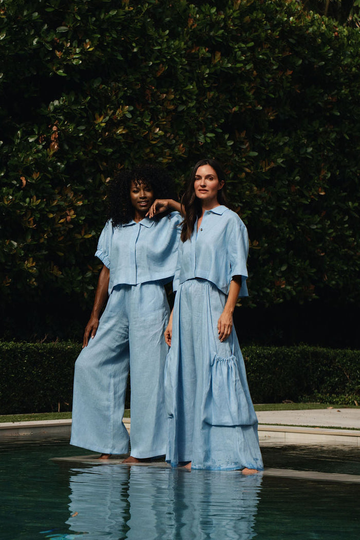 Two women standing in shallow pool water wearing 100% linen matching light blue short-sleeve jumpsuits or long dresses, with a dark green hedge background.