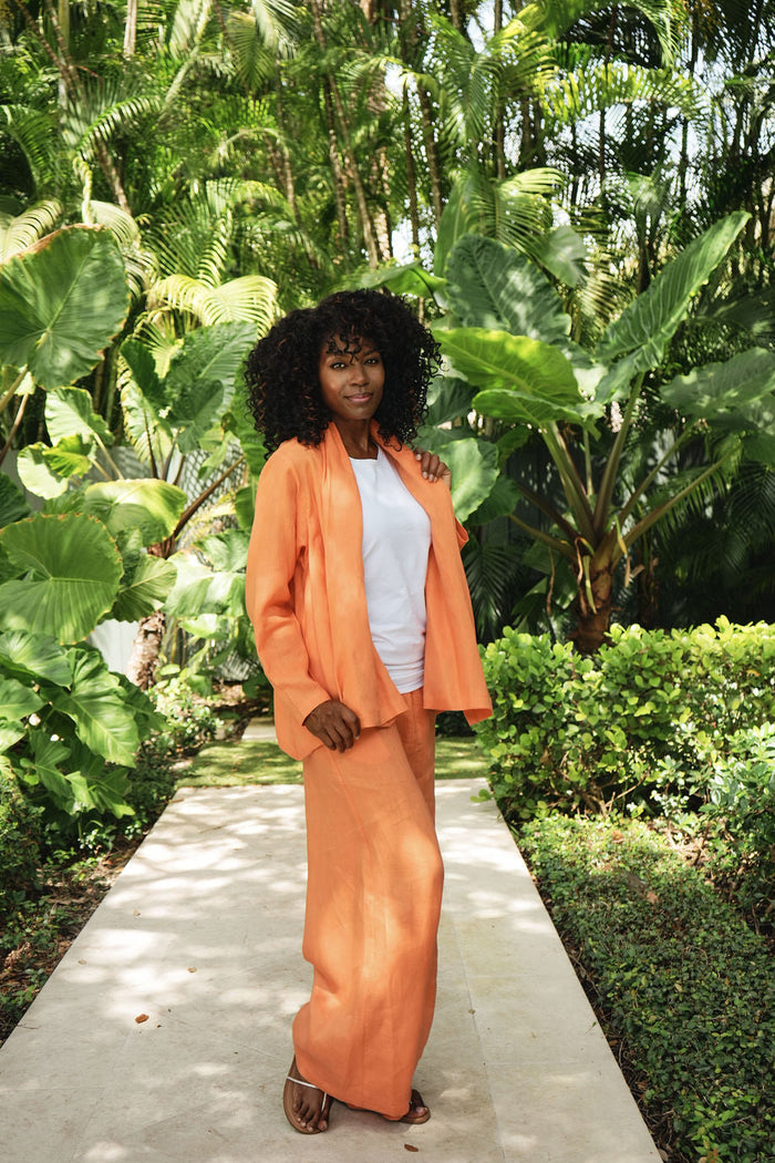 A woman stands on a concrete path surrounded by lush tropical green foliage, wearing a matching orange two-piece 100% linen outfit with a white top underneath.