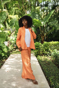 A woman stands on a concrete path surrounded by lush tropical greenery, wearing a bright orange 100% linen two-piece outfit with a white top and brown sandals.