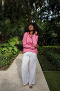 A woman stands on a stone pathway in a lush garden, wearing a bright pink long-sleeve top and 100% linen white wide-leg pants.
