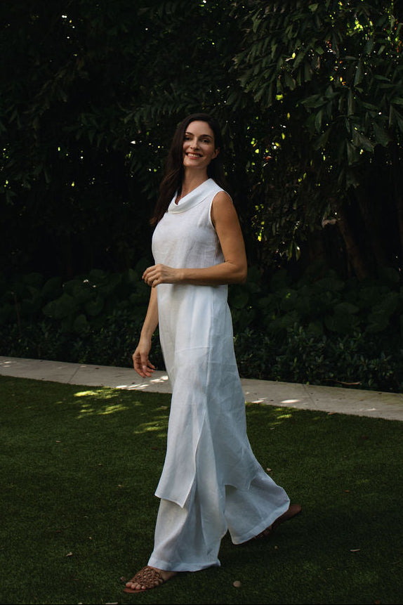 A woman wearing a 100% linen flowing white sleeveless dress walks across a grassy lawn in a garden with dense dark green foliage in the background.