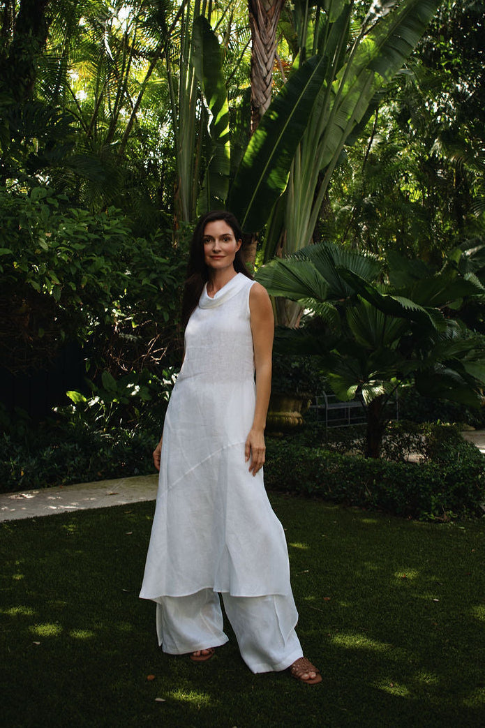 A woman wearing a 100% linen white sleeveless maxi dress stands in a lush tropical garden with dense green foliage and a stone path behind her.