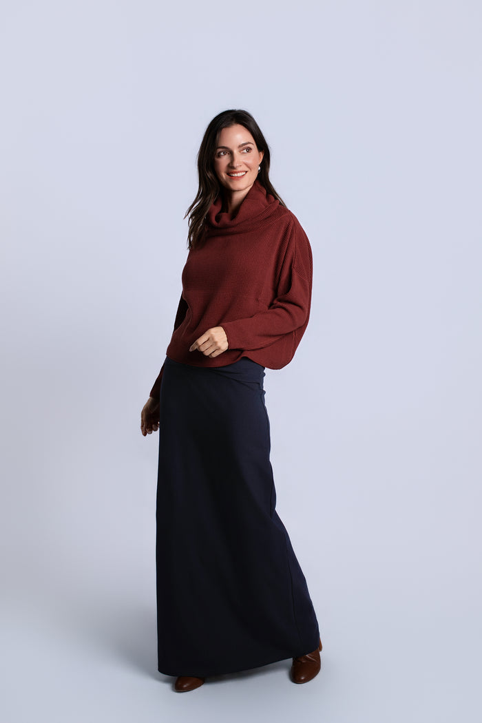 A woman modeling a dark red cowl-neck sweater and a long navy skirt against a light gray studio background.