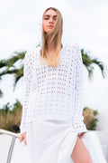 Woman in a beige patterned sweater and white pants standing on a beach at sunset.