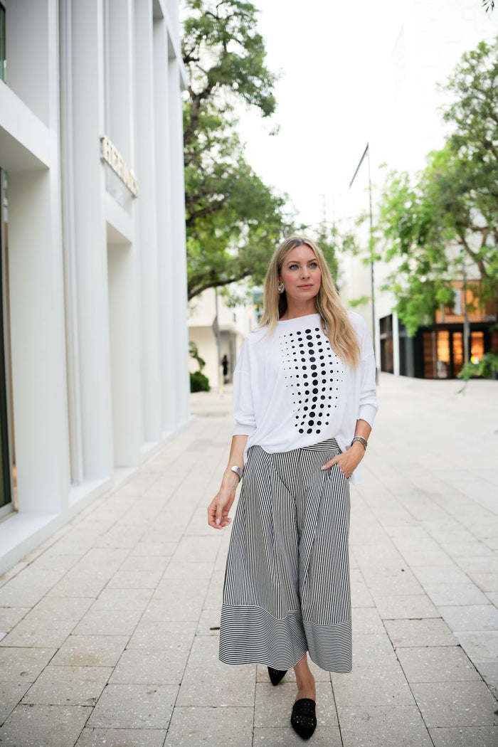 Woman wearing a white blouse with black pattern and gray wide-leg pants walking outdoors.