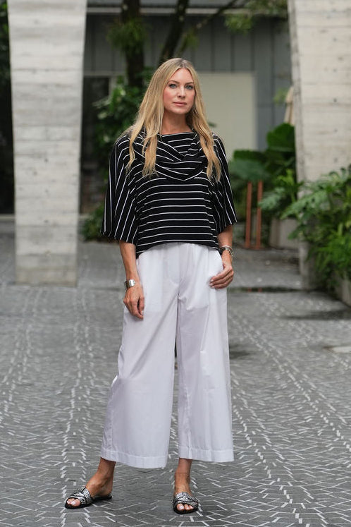 A woman stands outdoors wearing a black and white striped pima cotton top with wide-leg white cropped pants and dark flat sandals.