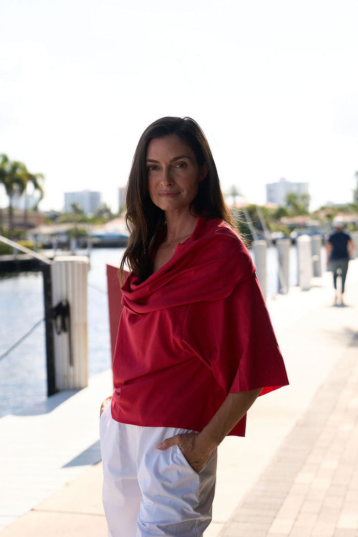 Woman in a red pima cotton jackie o top and white pants standing by a waterfront with trees and buildings in the background.
