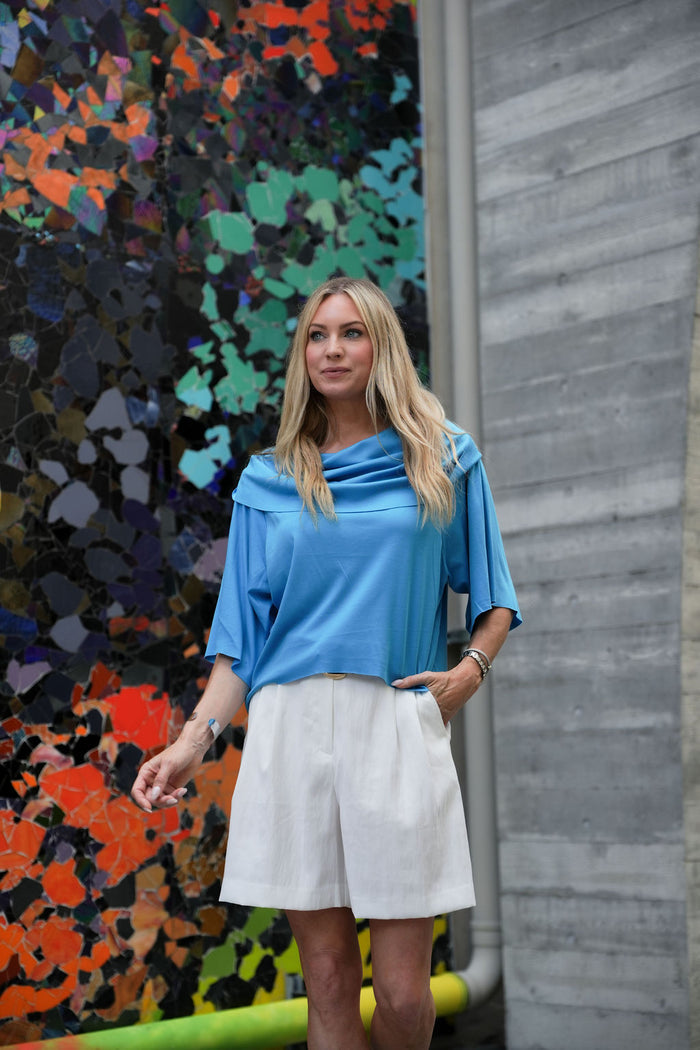 Woman wearing a blue pima cotton Jackie O top and white skirt standing in front of a colorful mural.