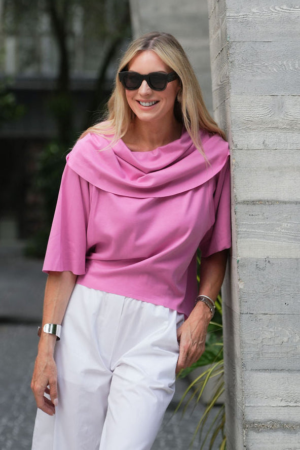 Woman in a pink pima cotton top and white pants standing against a gray concrete wall.
