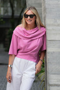 Woman in a pink pima cotton top and white pants standing against a gray concrete wall.