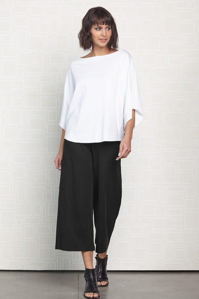 Woman wearing a white pima cotton top and black pants against a white wall.