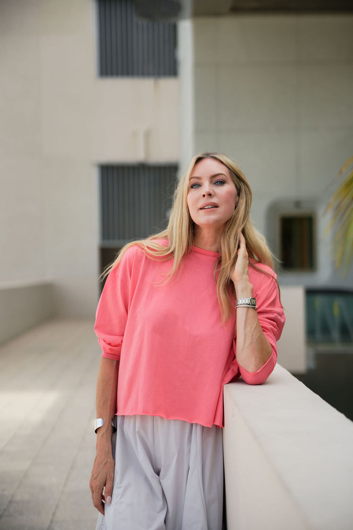 Woman in a coral pink pima cotton boxy t-shirt and gray pants standing in an indoor setting.