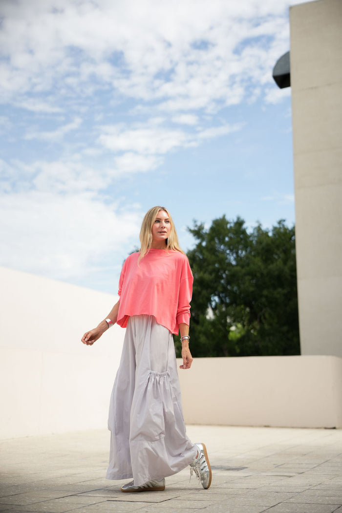 Woman in a coral pink pima cotton boxy t-shirt and light gray pants walking outdoors with a blue sky and trees in the background.
