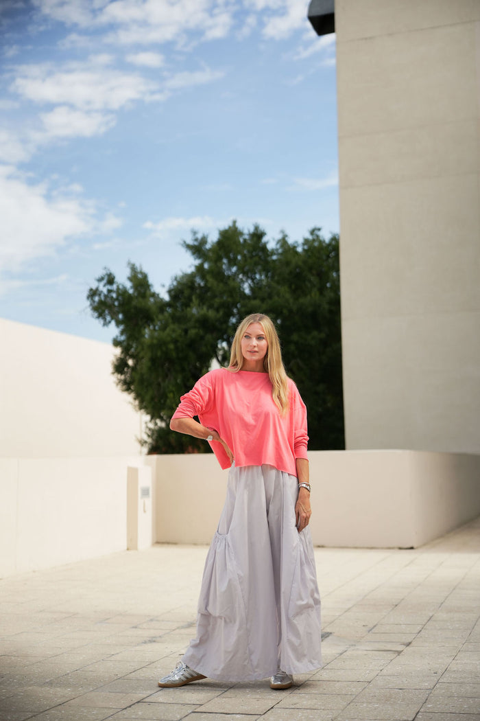 Woman in a coral pink pima cotton boxy t-shirt and light gray pants standing outdoors with a blue sky and tree in the background.