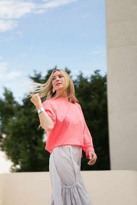 Woman in a coral pink pima cotton boxy t-shirt and gray pants standing outdoors with trees and blue sky in the background.
