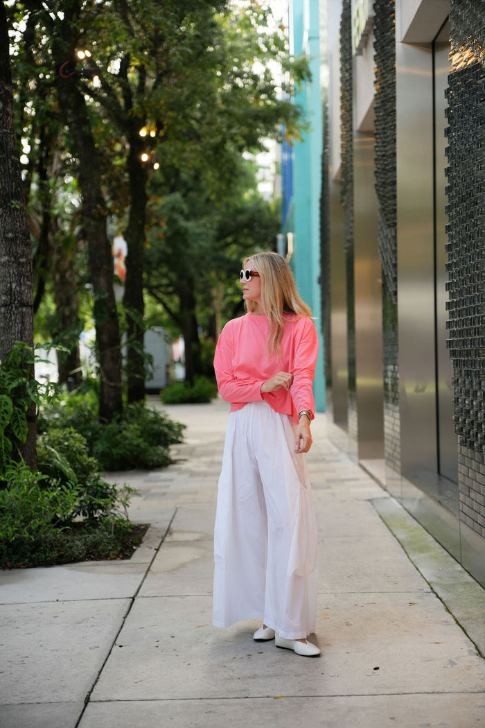 Woman in a coral pink pima cotton boxy t-shirt and white pants walking on a sidewalk with trees and modern architecture in the background.