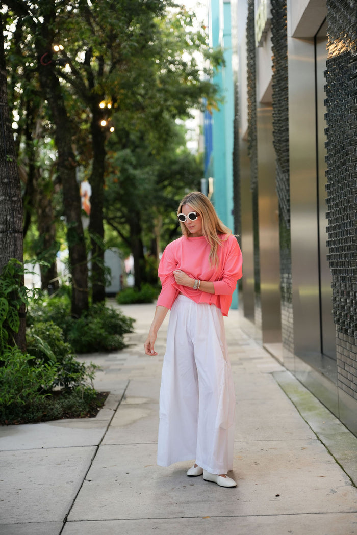Woman in a coral pink pima cotton boxy t-shirt and white pants walking outdoors near a modern building.