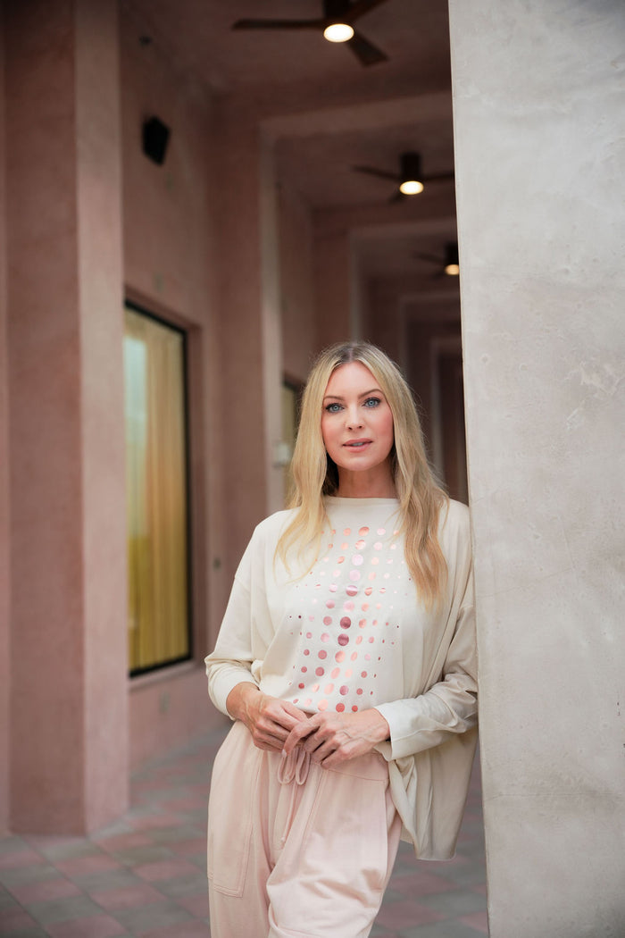 Woman standing in a hallway with pink walls and a painting on the wall wearing a stone and pink pima cotton boxy t-shirt with rose gold polka dots.