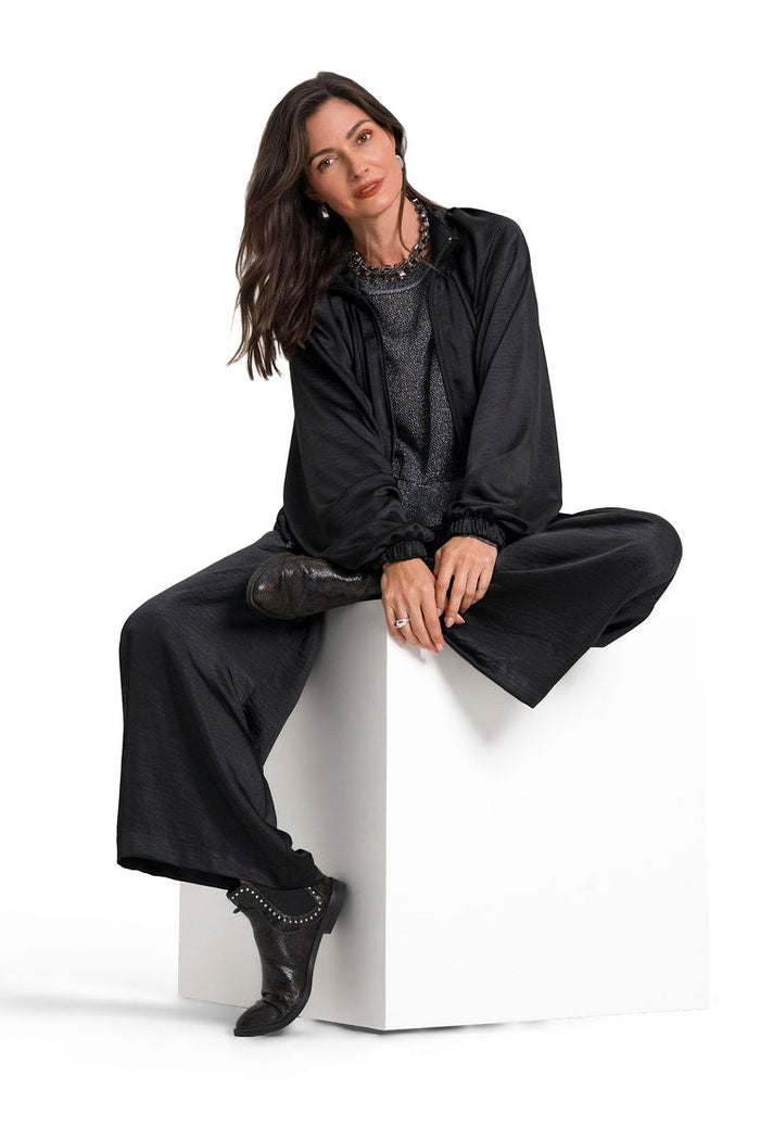A woman with long dark hair sits on a white cube pedestal against a solid white studio background. She is wearing a black wide-sleeve satin-like top, matching black wide-leg trousers, and black ankle boots with silver studs.